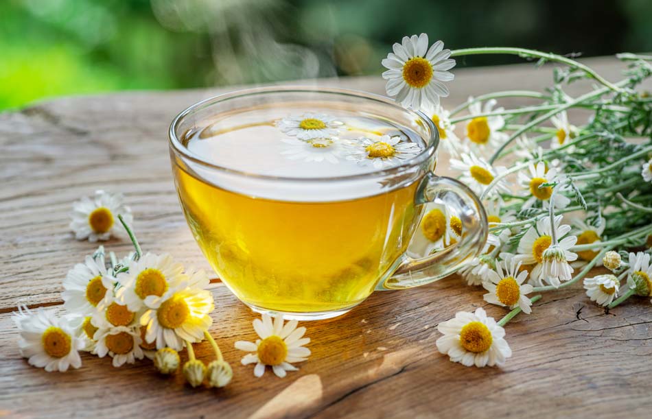 Clear glass mug filled with yellow tea surrounded by chamomile flowers on a wooden surface