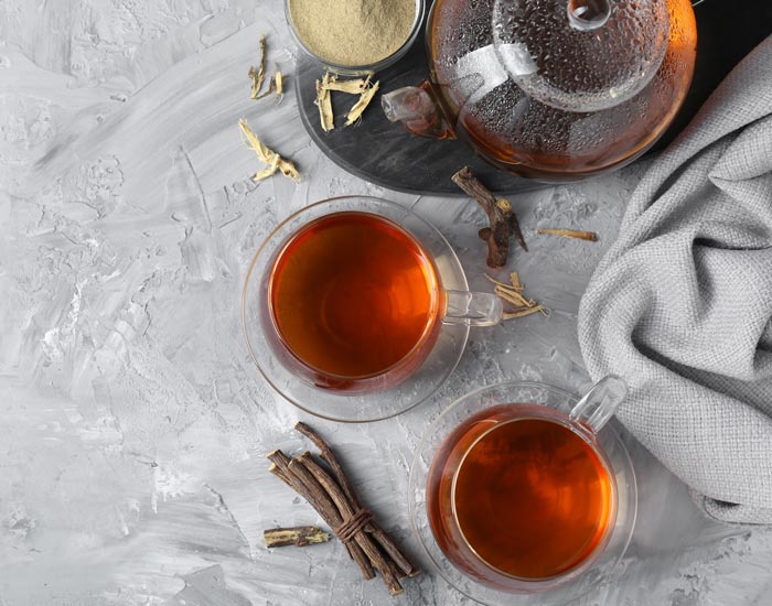 Two glass cups filled with tea on a gray surface with a teapot and licorice root.