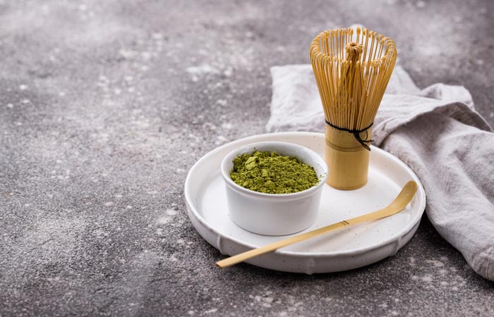 Matcha green tea powder in a small white bowl with a whisk and spoon on a gray surface.