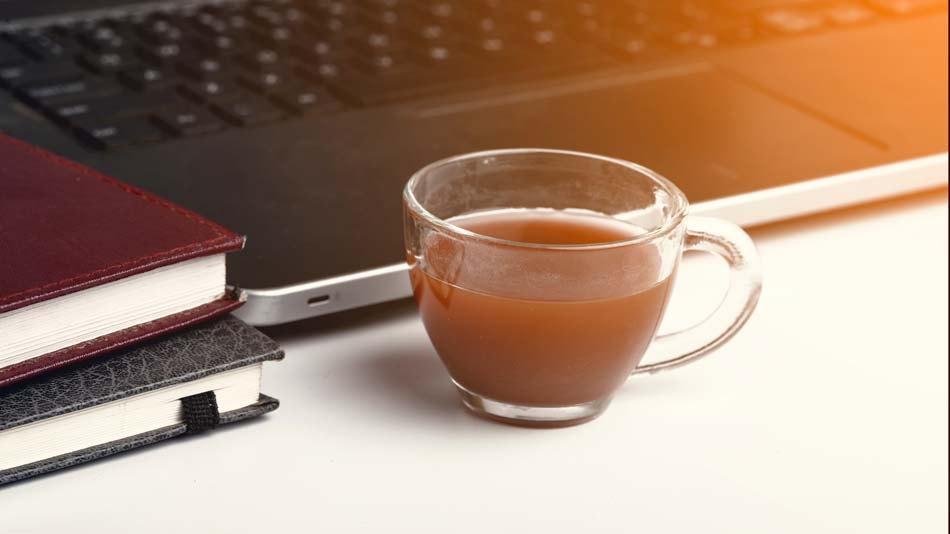 Glass mug of coffee on a desk with a laptop and notebooks.