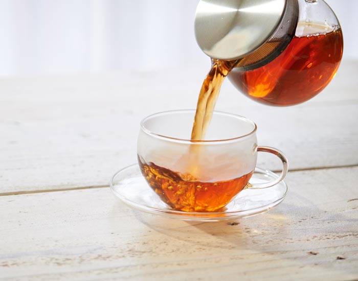 Tea being poured from a glass teapot into a clear glass cup on a wooden surface.