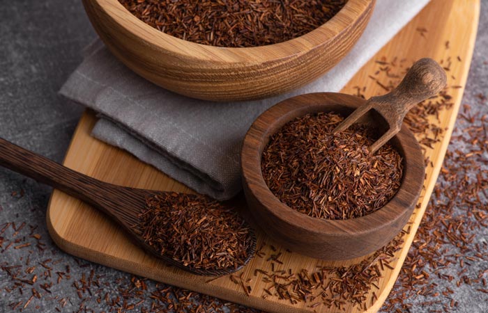 Wooden bowls and spoon filled with dark brown rooibos spice on a wooden board.