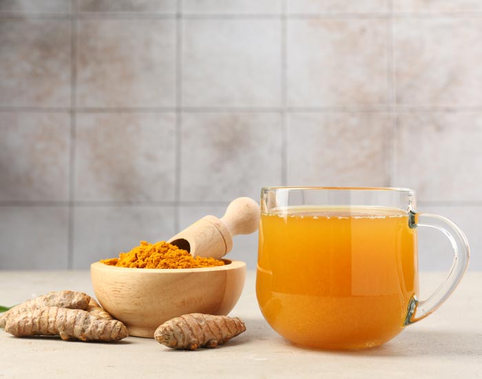 Glass mug of Turmeric Tea next to a wooden bowl of turmeric powder on a tiled surface.